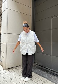 Pjaustytas šviesiai pilkas liemenė su apvaliais mygtukais, uždėta ant balto marškinio, derinama su tamsiai aptemptomis kelnėmis ir baltais sportbačiais. Po ja plokščias paviršius.