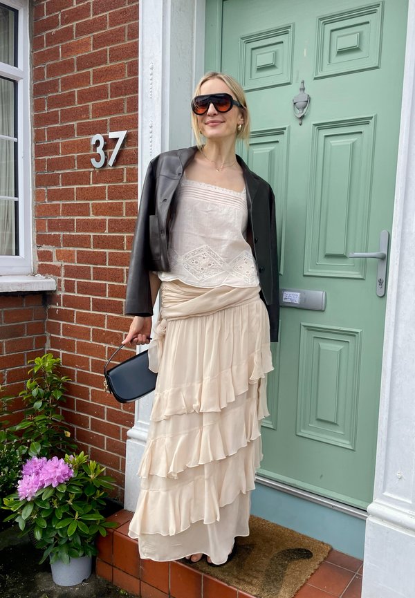 Vrouw met een beige gelaagde rok, witte kanten top, zwart leren jasje en grote zonnebril staat bij groene deur met nummer 37 met een zwarte handtas.