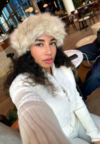 Un cappello in pelliccia sintetica di colore avorio, indossato con un gilet bianco con zip. Lo sfondo presenta mobili in legno e un'illuminazione soffusa.