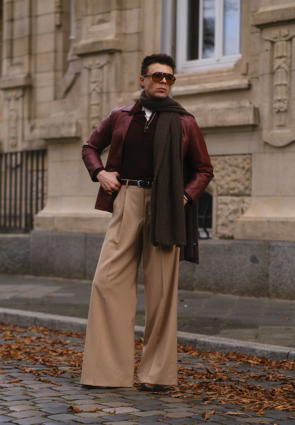 Bordeaux leren jack, bruine sjaal, donker shirt en beige wijde broek. Zonnebril maakt de outfit compleet, voor een stenen muur.