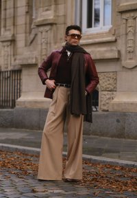 Bordeaux leren jack, bruine sjaal, donker shirt en beige wijde broek. Zonnebril maakt de outfit compleet, voor een stenen muur.