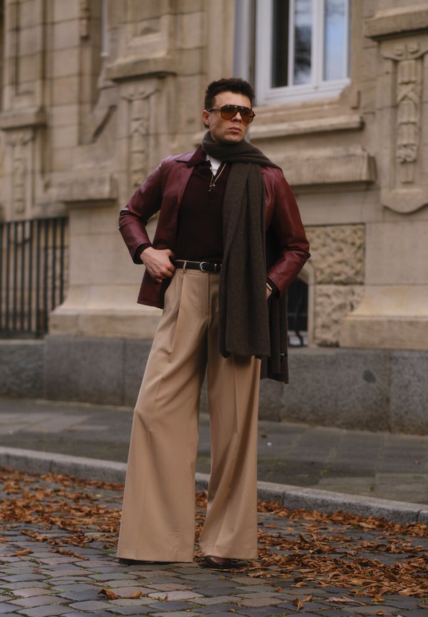 Bordeaux leren jack, bruine sjaal, donker shirt en beige wijde broek. Zonnebril maakt de outfit compleet, voor een stenen muur.
