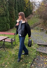 Crna kožna jakna s kremastim ovratnikom, sivi džemper, plave traperice i crna torbica. Model hoda travnatom stazom blizu zimzelenih stabala.