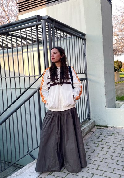 Chaqueta blanca con cremallera y rayas naranjas, combinada con una falda larga gris oscuro. De pie cerca de una valla metálica en una zona pavimentada.