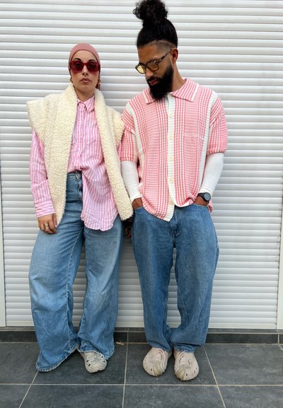 Dos personas están de pie frente a un fondo blanco. Una lleva una camisa de rayas rosas con un chaleco crema; la otra una camisa de cuadros rojos y blancos con mezclilla.