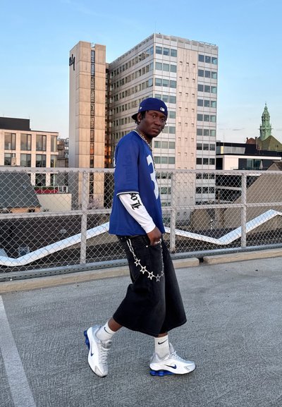 Jersey de malla azul con acentos blancos, pantalones de pierna ancha negros, detalle de cadena de estrellas plateadas y zapatillas Nike grises con acentos azules. Entorno urbano.