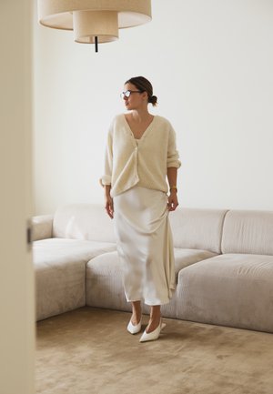 Cardigan de lana color crema con botones, falda de satén color crema y tacones blancos de punta, de pie sobre una alfombra marrón claro junto a un sofá beige.