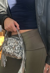 Sac à main en sequins argentés avec des accents de franges, présentant une forme structurée et des sangles métalliques, tenu par une personne portant une tenue verte olive.
