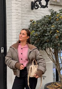 Femme portant une veste marron et un pull rose, tenant un sac à main beige, debout près d'un mur en briques blanches et d'un arbre feuillu en plein air.