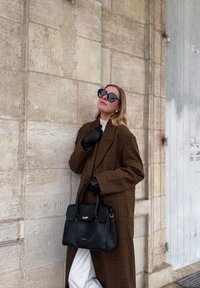 Femme portant un manteau marron à carreaux, des gants noirs et de grandes lunettes de soleil, tenant un sac à main noir, debout contre un mur de pierre beige.