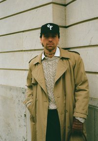 Beige trenchcoat över en kabelstickad tröja, svart keps med vit logotyp, mörka byxor, bruna läderhandskar, stående mot en stenmur.