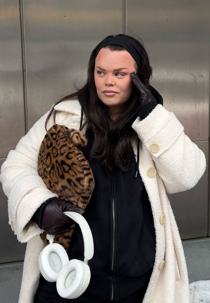Femme portant un manteau blanc et un bandeau noir, tenant un casque blanc et un sac à imprimé léopard, touchant sa tempe avec une main gantée.