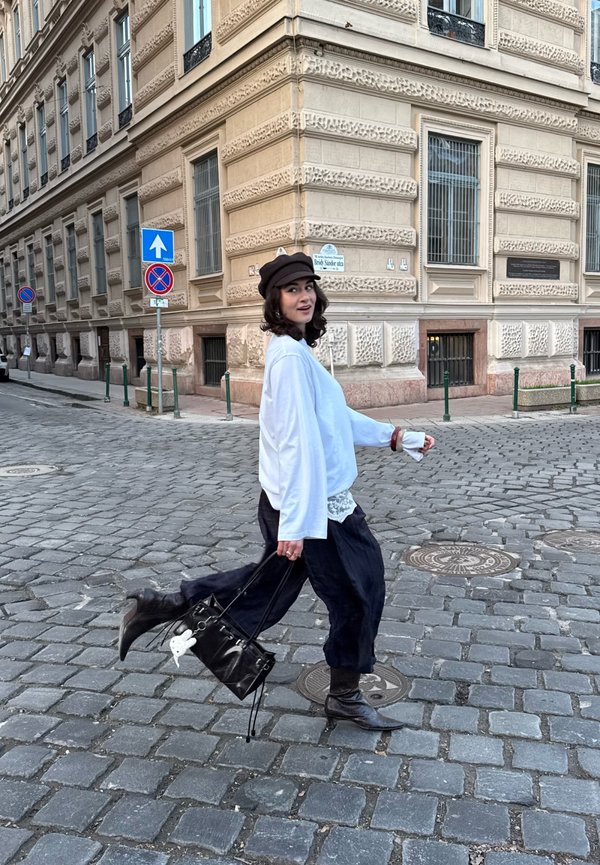 Fiatal nő fehér pulóverben, fekete nadrágban és kalapban, aki köves utcán sétál egy díszes bézs épület mellett, fekete kézitáskát hordva.