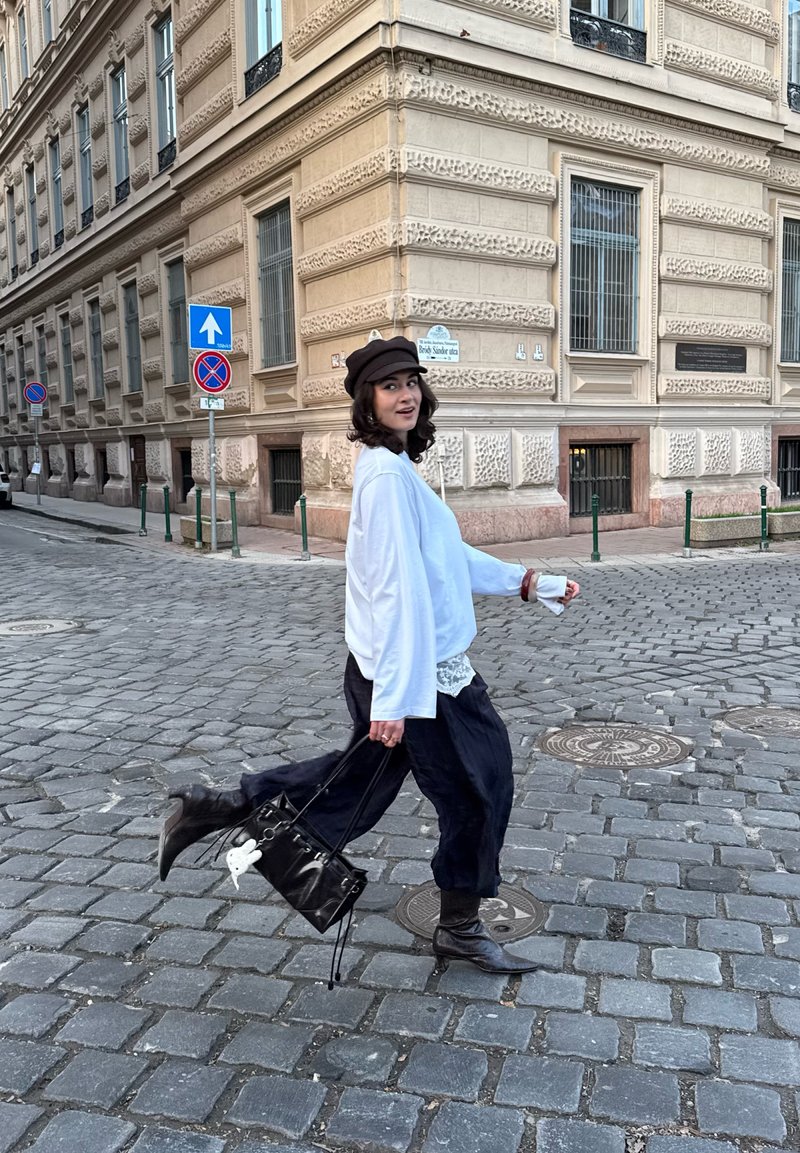 Giovane donna con maglione bianco, pantaloni neri e cappello che attraversa una strada lastricata vicino a un edificio beige ornato, portando una borsa nera.