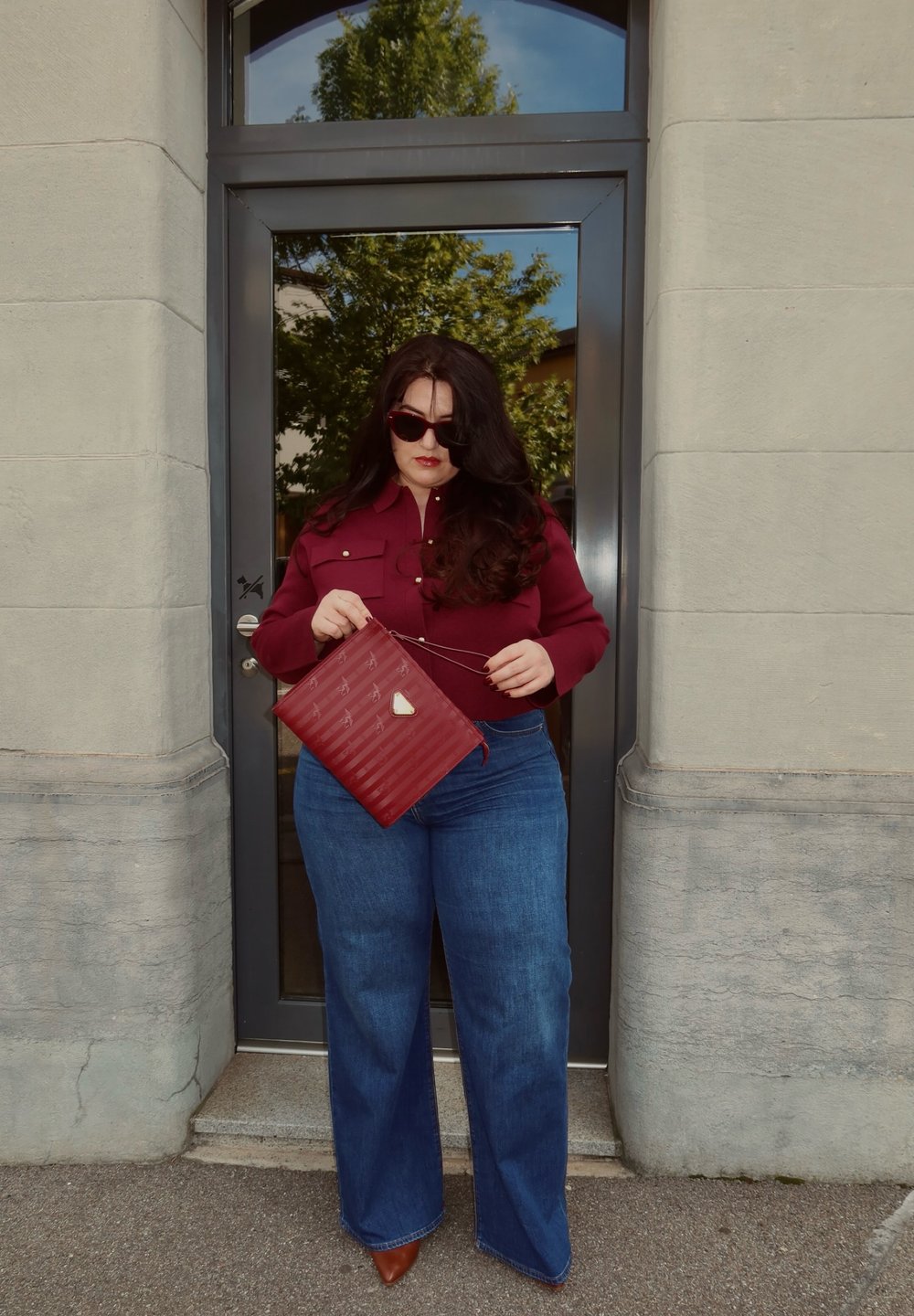 Mujer con cabello largo y oscuro que lleva gafas de sol, blusa burdeos, jeans de pierna ancha azules y zapatos marrones, sosteniendo un bolso rojo frente a una puerta de vidrio.
