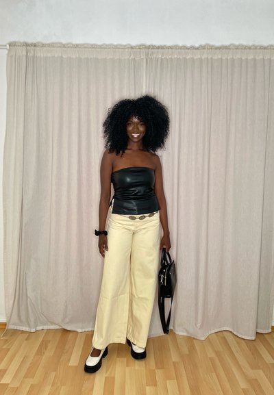 Top strapless de cuero negro, pantalones de pierna ancha crema con un cinturón decorativo, bolso negro y zapatos de plataforma en blanco y negro sobre un suelo de madera.