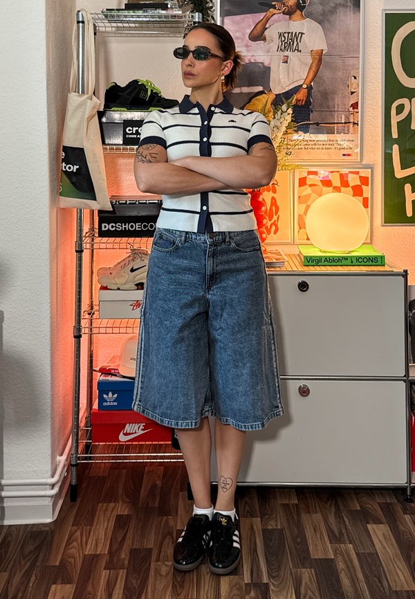 Persoon met zonnebril, gestreept shirt en denim shorts staat met gekruiste armen in een kamer met sneakers en kleurrijke posters achter zich.