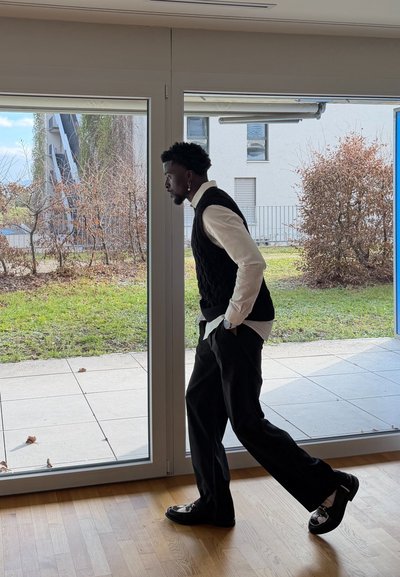 Hombre con chaleco negro y camisa blanca caminando en el interior cerca de grandes puertas de vidrio que dan a un jardín con arbustos secos y verdes.