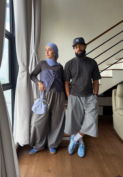 Mujer con hijab, chaleco, pantalones anchos sosteniendo una bolsa, y hombre con barba, gorra, camiseta de gran tamaño y pantalones cortos, de pie en el interior junto a una ventana y una escalera.