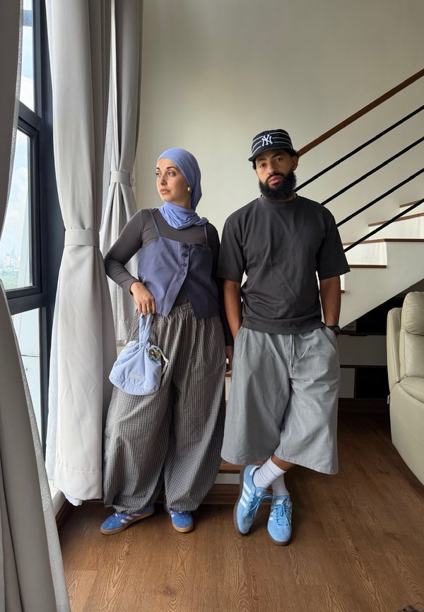 Kvinne i hijab, vest, vide bukser som holder en veske, og mann med skjegg, lue, baggy t-skjorte og shorts som står innendørs ved et vindu og en trapp.