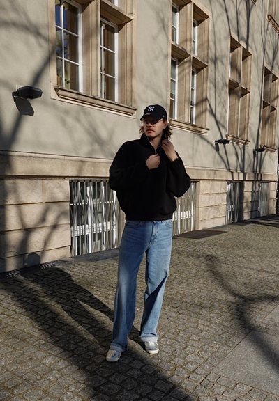 Joven con chaqueta negra, vaqueros azules y gorra de los New York Yankees, de pie en una calle adoquinada frente a un edificio con múltiples ventanas.
