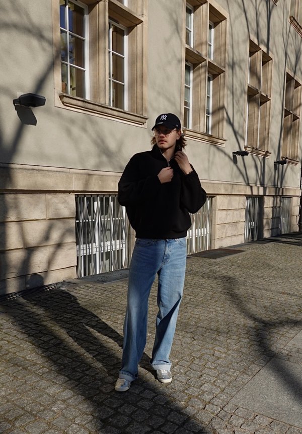 Ung mann iført svart jakke, blå jeans og New York Yankees-caps står på brosteinsgate foran en bygning med flere vinduer.