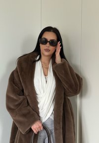 Manteau en fausse fourrure marron, haut cache-cœur blanc, pantalon gris. Accessoires comprenant des lunettes de soleil oversize et un sac à main marron. Textures lisses et design drapé.