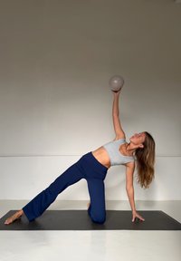 Vêtements de sport comprenant un débardeur ajusté gris clair et un pantalon de yoga bleu foncé. La personne effectue une étirement latéral sur un tapis noir, en levant une balle pâle.