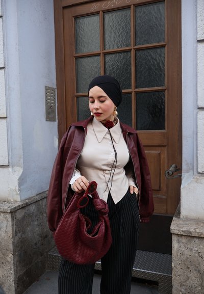 Chaqueta de cuero marrón, camisa crema de botones, pantalones negros a rayas. Sosteniendo un bolso tejido marrón y gafas de sol rojas. Pañuelo negro en la cabeza.