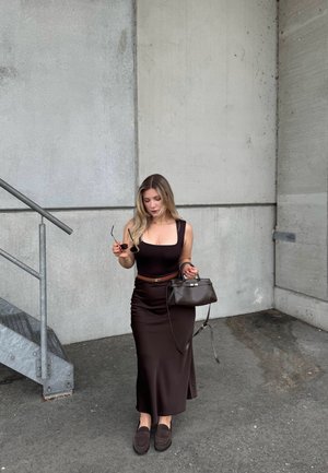 Mujer con un vestido marrón oscuro que sostiene unas gafas de sol y un bolso a juego, de pie cerca de unas escaleras de metal contra una pared de hormigón.