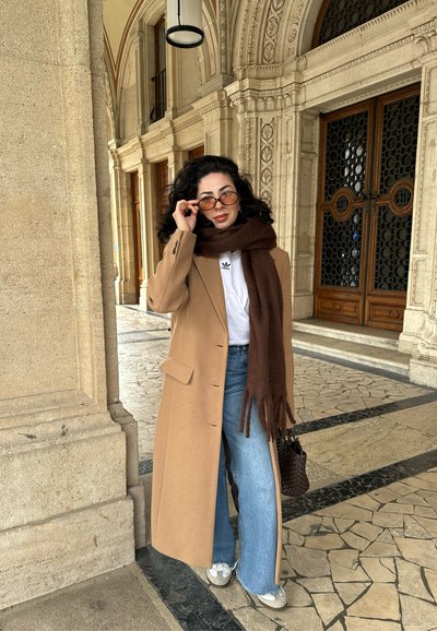 Abrigo de lana beige, bufanda marrón, camiseta blanca y vaqueros de pierna ancha de color azul. Los accesorios incluyen gafas de sol y un pequeño bolso de palma.