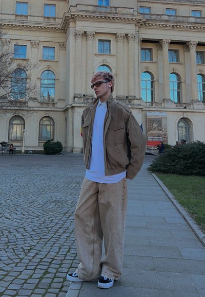 Joven con gafas de sol y auriculares, vistiendo pantalones beige de gran tamaño, chaqueta marrón y camisa blanca, de pie en una plaza pavimentada frente a un gran edificio histórico.