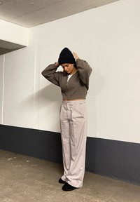 Brązowa krótka dzianinowa kurtka, biała koszulka, jasnobeżowe spodnie, czapka beanie w kolorze czarnym i czarne obuwie w minimalistycznym wnętrzu.