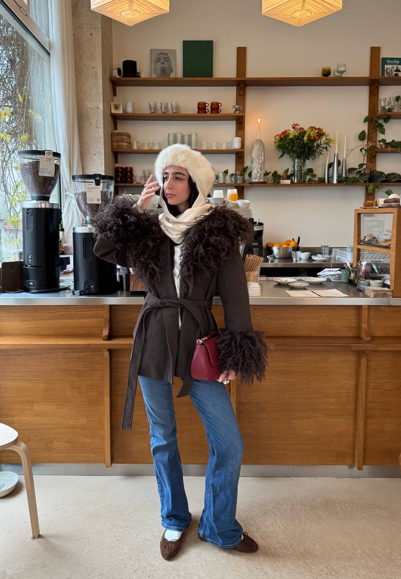Vrouw met een pluizige witte hoed, een bruine jas met bontkappen, blauwe jeans en een rode handtas staat in een gezellige café bij koffiemachines.
