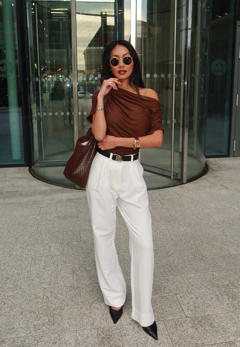 Brown off-the-shoulder top, high-waisted white trousers, black pointed heels, and brown textured tote bag. Accessories include a watch and sunglasses.