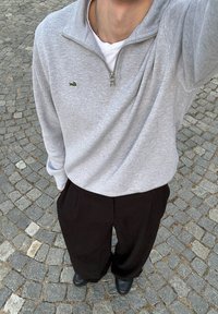 Grå sweatshirtsjakke med lynlås og ribbet krave samt lille grønt logo, parret med løstsiddende sorte bukser og sorte sko, stående på stenhåndværk.