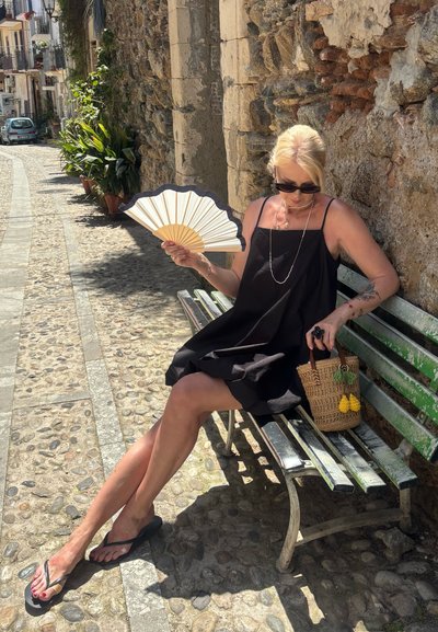 Vestido negro sin mangas, bolso de mimbre con acentos amarillos, abanico decorativo y chanclas negras; sentado en un banco de madera verde, camino de piedra cercano.