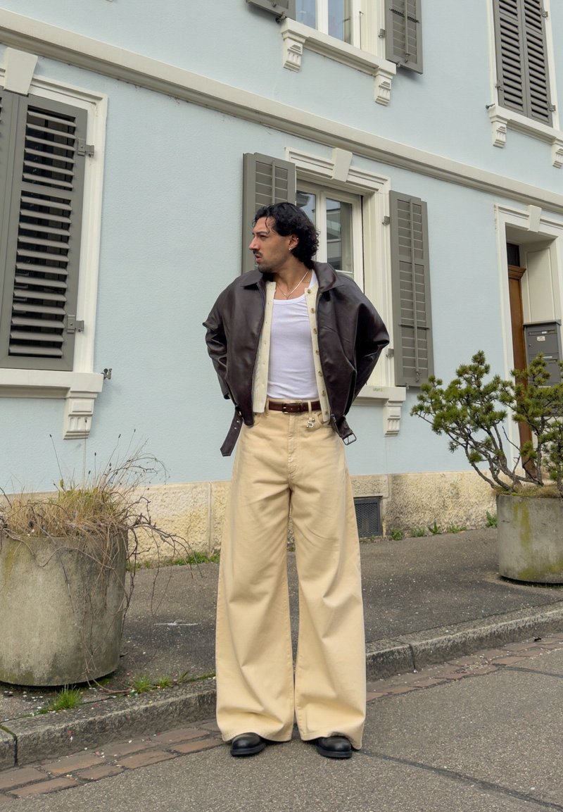 Homme portant un pantalon large beige, un débardeur blanc, un cardigan crème et une veste en cuir marron foncé, debout dans la rue près d'un bâtiment bleu pastel.