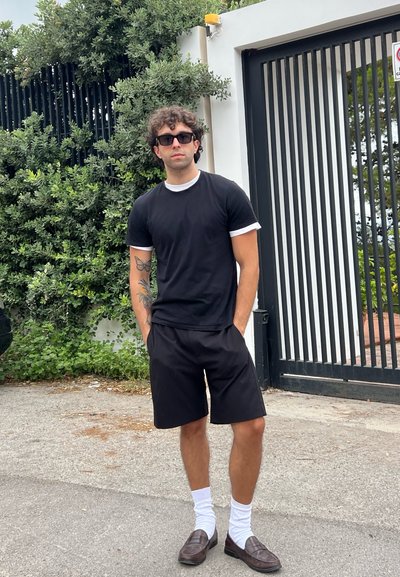 Camiseta negra con cuello blanco, pantalones cortos negros, calcetas blancas y mocasines marrones. De pie contra un fondo de vegetación y una entrada enrejada negra.