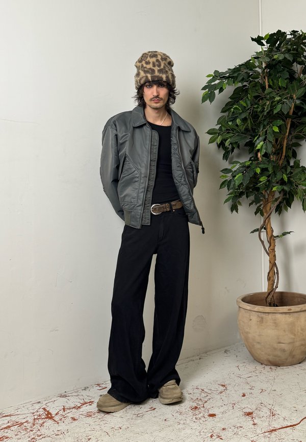 Jeune personne portant une veste grise, une chemise noire et un pantalon large noir, debout les mains derrière le dos près d'une plante en pot à feuilles.