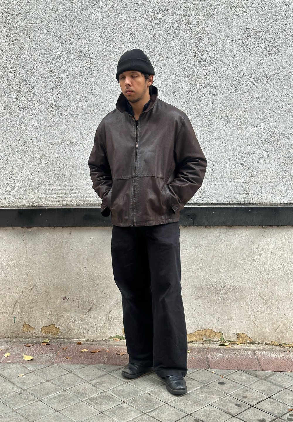Brown leather jacket with a high collar, black beanie, and loose black trousers, standing against a light grey wall. Shoes are black.
