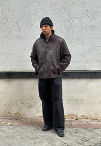 Brown leather jacket with a high collar, black beanie, and loose black trousers, standing against a light grey wall. Shoes are black.