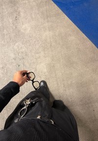 Lunettes rectangulaires noires tenues à la main, portées avec un haut noir texturé et un pantalon noir ample. Le sol présente un revêtement gris avec des accents bleus.