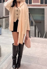 Cardigan in maglia crema sopra un top beige con bottoni neri, abbinato a una mini gonna nera e stivali alti lucidi neri. Borsa rosa.