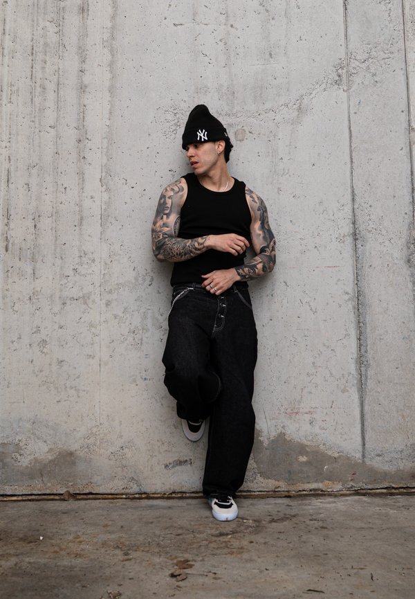 Mladý muž s tetováním na celé paži, oblečený v černém tílku, volných černých džínách, teniskách a černé čepici New York Yankees, opírající se o betonovou zeď.