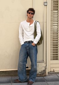 Weißes langärmliges Hemd mit Henley-Ausschnitt, lockere blaue Jeans, braune Schuhe und eine olivgrüne Schultertasche; Sonnenbrille vervollständigt den Look.