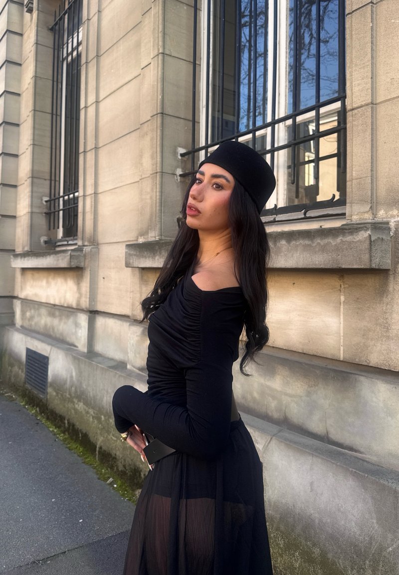 Vrouw met lang donker haar, gekleed in een zwarte off-shoulder top, doorschijnende zwarte rok en zwarte hoed, staand bij een stenen gebouw met hoge ramen.