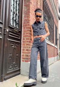 Czarna dżinsowa kamizelka z krótkim krojem i zatrzaskami, zestawiona z szerokimi czarnymi dżinsami. Obuwie to masywne białe botki. Tło to czerwona cegła.