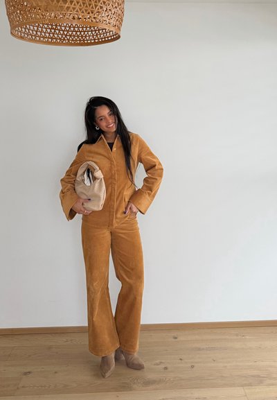 Mujer con traje de pana color beige y botas beige, sosteniendo un bolso fruncido beige, de pie sobre un suelo de madera bajo una pantalla de lámpara tejida.