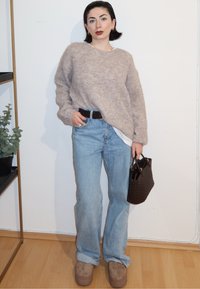 Femme appuyée contre un mur, portant un pull beige, un jean bleu clair, des chaussures marron, et tenant un sac à main tressé marron foncé.